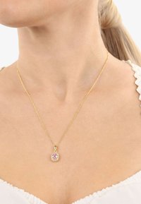 Guldkedja med en känslig kedja som har en oval rosa hänge prydd med små klara stenar, bärs nära halslinjen.