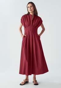Červené maxi šaty s krátkymi rukávmi, vysokým golierom, skladaným pásom a bočnými vreckami. Hladká tkanina s voľným strihom a plynulou sukňou.