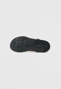 Semelle en caoutchouc noire avec des motifs de traction texturés, forme arrondie et légère courbure. Bords renforcés et petit accent rouge près du talon.