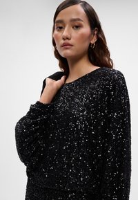 Juodas blizgučiais puoštas megztinis su laisvu siluetu, apvaliu iškirpimu ir ilgomis rankovėmis; blizgus tekstūros paviršius suteikia vizualinį sužavėjimą. Modelis laiko juodą švarką.