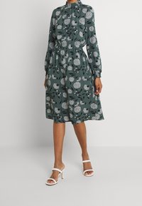 Robe fleurie verte avec un col montant, des manches longues, une taille élastique et une jupe à hauteur des genoux. Présente des motifs floraux blancs et noirs.