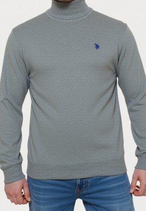 Mann trägt einen hellgrauen Rollkragenpullover mit blauem, aufgesticktem Logo und blaue Jeans, steht vor einem einfarbigen Hintergrund.