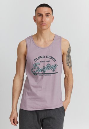 Homme aux cheveux courts portant un débardeur lilas avec le texte « Blend Denim Surfing » et un tatouage sur la partie supérieure de son bras gauche.