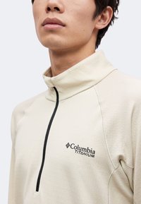 Άνδρας που φοράει ένα ανοιχτό μπεζ Columbia Titanium πουλόβερ με φερμουάρ έως το στήθος και με υφή στο ύφασμα.