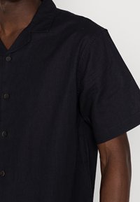 Kurzärmliges, knöpfbares Hemd aus dunkelblauem Stoff mit subtiler Struktur, ausgestattet mit einem Reverskragen und schlichten braunen Knöpfen.