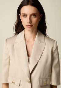 Joven con cabello largo y oscuro lleva un blazer beige con botones dorados en forma de abeja contra un fondo beige liso.