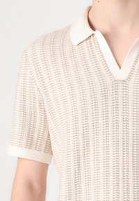Svijetlo bež pleteni polo majica s V-izrezom, teksturiranim uzorkom, kratkim rukavima i bijelim rebrastim ovratnikom.