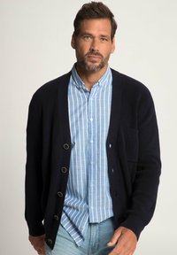 Cardigan lavorato a maglia di colore blu navy con grandi bottoni rotondi in bronzo, indossato sopra una camicia button-up a righe blu chiaro. Presenta una tasca frontale e una texture a coste.