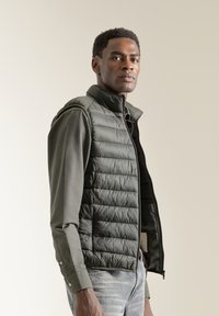 Chaleco acolchado verde con cuello alto y cremallera frontal, usado sobre una camisa de manga larga, que presenta una superficie texturizada y costuras horizontales.