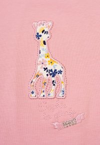 Tessuto rosa con una silhouette di giraffa ricamata con motivi floreali, accentuata da cuciture rosa e verdi e il testo "Sophie la girafe Paris."