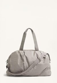 Harmaa kangasduffeli, jossa on sileä pinta, kaksinkertaiset kahvat ja irrotettava olkahihna. Pyöristetty muotoilu vetoketjullisella suljennalla.