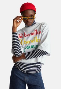 Bílý oversized tričko s barevným nápisem "benetton", vrstvené přes tmavě modrý a bílý pruhovaný rolák. Kombinováno s tmavými džínami a červenou čepicí.