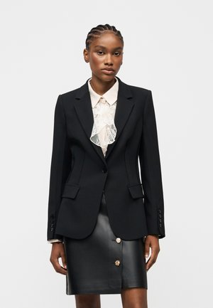 Žena s copánky, která má na sobě černý blazer přes bílou halenku s volánky a černou koženou sukni s ozdobnými knoflíky.