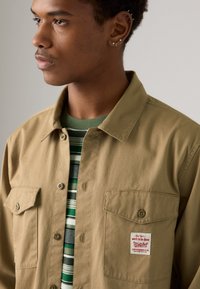 Tan button-up shirt met twee borstzakken en een rood logo patch, bovenop een groen-wit gestreept T-shirt. Soepele katoen stof.