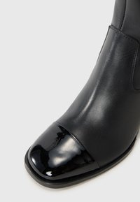 Bottines noires en cuir avec un bout en vernis noir brillant, texture lisse et une silhouette épurée. Équipées de panneaux latéraux élastiques pour un enfilage facile.