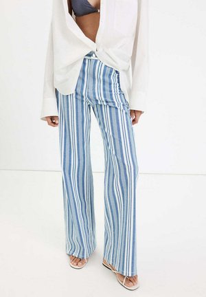 Femme portant un pantalon taille haute rayé vertical bleu et blanc, un chemisier blanc ouvert, un soutien-gorge noir et des sandales blanches sur un fond blanc.