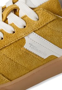 Baskets en daim jaune avec des accents en cuir blanc et des lacets blancs. Présente des détails cousus et une semelle en caoutchouc de teinte brun clair. Logo de la marque visible.
