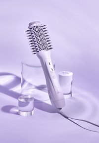Sèche-cheveux rond avec brosse, de couleur blanche, doté d'une tête à poils texturés, d'un bouton de contrôle et d'un cordon d'alimentation, sur un fond violet doux.