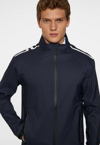 Ung man iklädd en marinblå zip-up-jacka med hög krage och vita randdetaljer på axlarna, stående mot en enfärgad bakgrund.