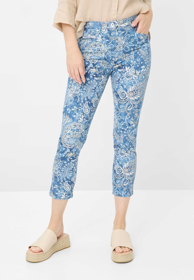 Jeans bleus à motifs avec un design floral paisley, longueur cropped, poches avant et silhouette ajustée. Portés avec des sandales à plateforme beiges.