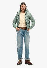 Chaqueta de abrigo acolchada verde menta con capucha, suéter de punto beige de trenzas, jeans de color azul claro y botas marrones de cordones. Conjunto casual y en capas.
