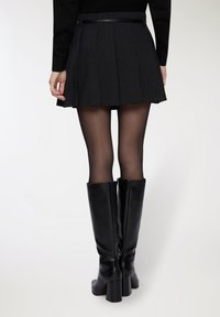 Donna che indossa una mini gonna nera a righe, collant neri trasparenti e stivali neri con tacco alti fino al ginocchio, vista da dietro.