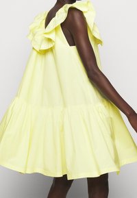Personne à la peau foncée portant une robe jaune ample sans manches avec des épaules volantées et une jupe évasée sur un fond uni.