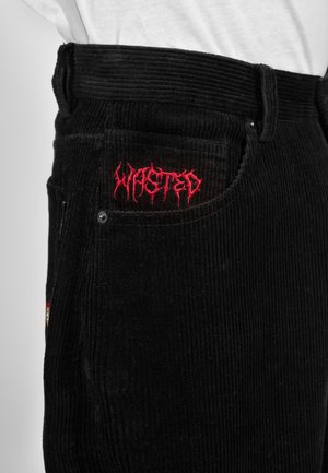 Schwarze Cordhosen mit rot besticktem Schriftzug „WASTED“ auf der kleinen vorderen Tasche, getragen mit einem in das Hosenbein gesteckten weißen Hemd.