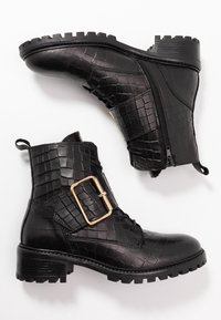 Botines negros de textura con tacones gruesos, cremallera lateral, cordones y grandes correas decorativas de hebilla dorada sobre un fondo blanco.