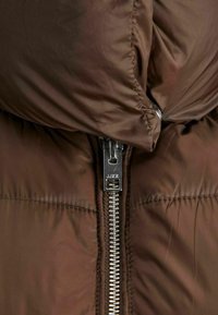 Veste rembourrée marron avec une texture lisse et brillante. Présente une fermeture éclair argentée et un col haut, soulignant la chaleur et l'isolation.