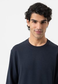 Jonge man met krullend donker haar, gekleed in een marineblauw longsleeve shirt, kijkt met een lichte glimlach naar de camera, op een effen witte achtergrond.