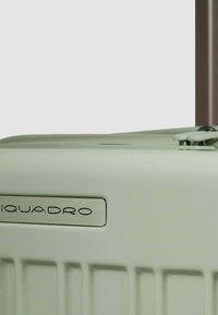 Maleta dura verde claro con ranuras verticales, una placa de metal plateada, y un mango retráctil sobre un fondo marrón.