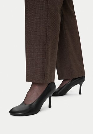 Kobieta ubrana w brązowe spodnie w kratę, czarne prześwitujące rajstopy i czarne skórzane czółenka na średnim obcasie, stojąca na białym tle.