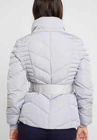 Femme vue de dos portant une veste matelassée gris clair avec un col montant et une large ceinture portant l'inscription "GUESS".