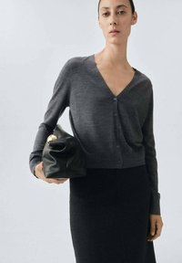 Dunkelgrauer Cardigan mit V-Ausschnitt, Knopfverschluss, langen Ärmeln und kurzem Schnitt. Kombiniert mit einer schwarzen Ledertasche.