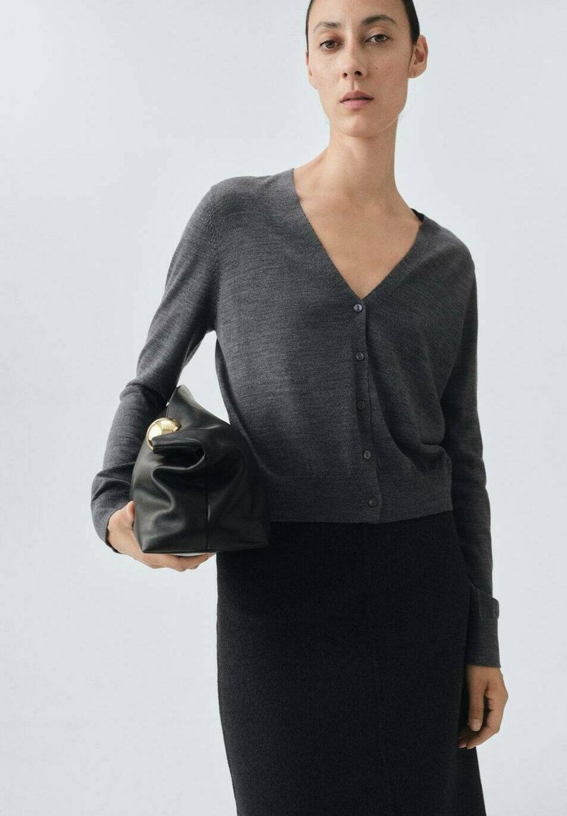 Dunkelgrauer Cardigan mit V-Ausschnitt, Knopfverschluss, langen Ärmeln und kurzem Schnitt. Kombiniert mit einer schwarzen Ledertasche.