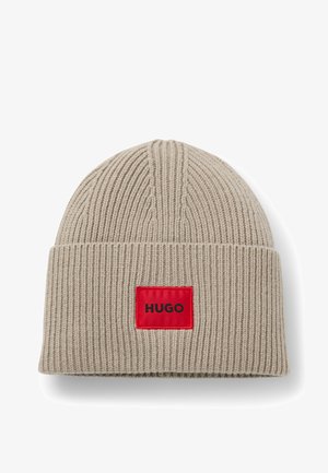 Béret en tricot beige clair avec une texture côtelée. Il présente un patch rectangulaire rouge avec le texte "HUGO" en noir à l'avant.