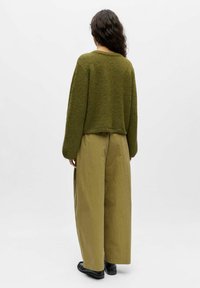 Vihreä neulotusta langasta valmistettu lyhyt neule, leveät hihat, yhdistetty oliivinvihreisiin leveälahkeisiin housuihin ja mustiin jalkineisiin, kuvattu takaa.