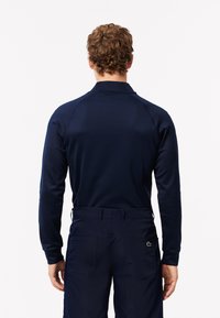 Polo de manga larga azul marino con cuello alzado, tejido texturizado y un acento de logo verde en la parte baja de la espalda.