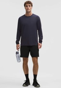 Granatowa sportowa koszulka z długim rękawem o fakturowanej tkaninie, czarne spodenki oraz czarne trampki; trzyma przezroczystą butelkę na wodę z pokrywką.