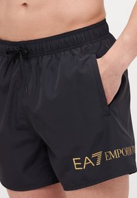 Pantaloni scurți de baie negri, din material ușor, cu talie elastică, șnur de ajustare, buzunare laterale și detalii cu un logo auriu.