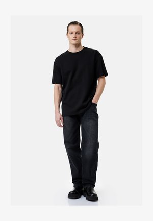 Man standing with one hand in pocket, wearing black oversized t-shirt, dark jeans, and black leather boots on a plain light background.