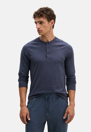 Man draagt een donkerblauw henleyshirt met lange mouwen en drie knopen en donkerblauwe broek met trekkoord, kijkt naar de zijkant.