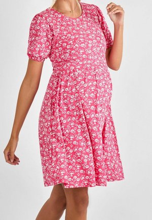 Vrouw in een roze bloemenjurk tot de knie, met korte pofmouwen en een gouden hartketting, met één hand omhoog en de andere tegen haar zij.