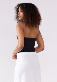 Sort strapless top, tætsiddende design, parret med en hvid omslagsskørt. Naturlige krøller rammer profilen, sat mod en lys baggrund.