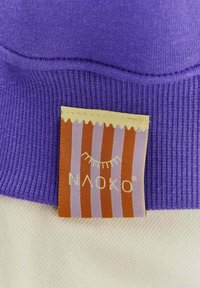 Fialová žebrovaná látka s visačkou s oranžovými a zlatými pruhy. Na visačce je napsáno "NAOKO" zlatým písmem a je zde grafika slunce.