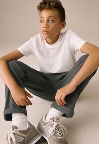 T-shirt en coton blanc, pantalon gris foncé, revers retroussés, baskets grises avec des accents et lacets blancs, assis sur un fond clair.