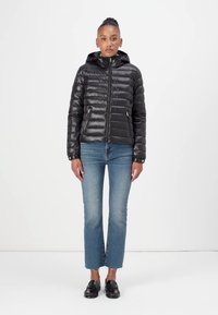Zwarte pufferjack met horizontale quiltpatronen, rits aan de voorkant en capuchon. Gecombineerd met lichtblauwe flared jeans en zwarte instapschoenen. Soepele textuur.