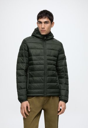 Homme portant une veste matelassée à capuche vert foncé avec fermeture éclair à l'avant et un pantalon marron, debout devant un fond clair uni.