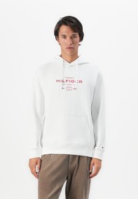 Homme portant un hoodie blanc Tommy Hilfiger avec poche kangourou et un pantalon en velours côtelé marron, se tenant contre un fond blanc uni.